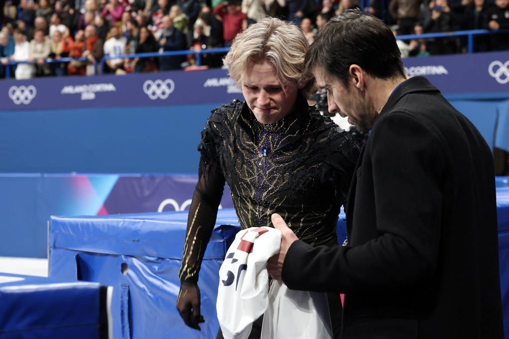 A male figure skater with blonde hair in a black and gold costume looks down, while a man in a black coat holds a white towel for him.