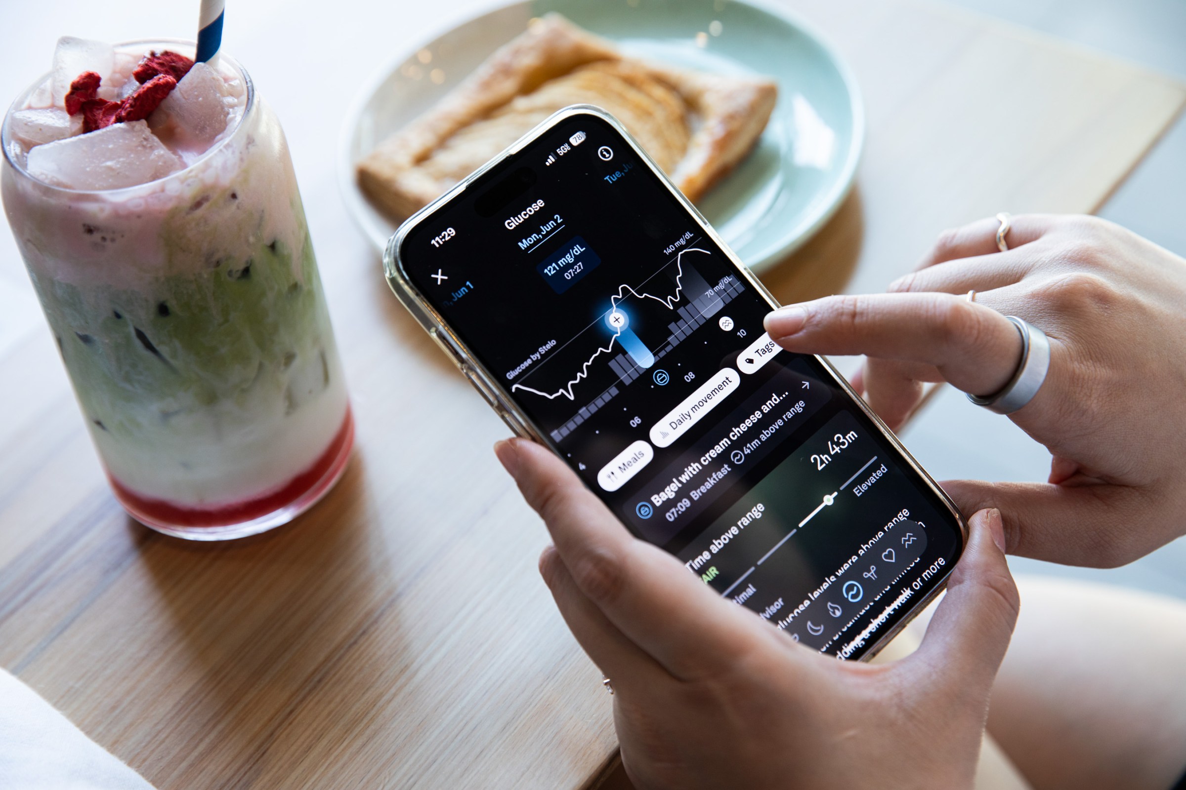 Person wearing a silver Oura Ring 4 looks at glucose readings with a strawberry matcha latte and a pastry seen on a wooden table in the background. 