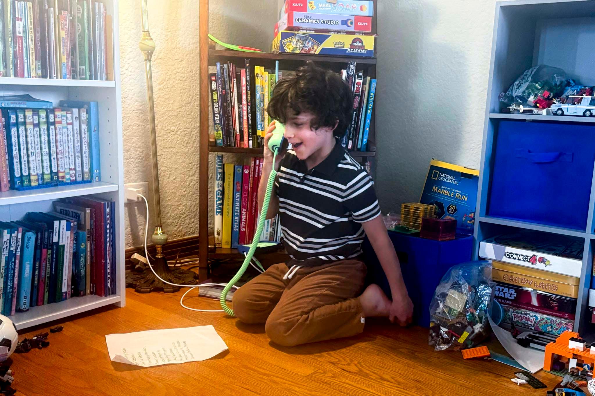 A kid in a striped polo sits on his knees on a wooden floor, surrounded by toys and books, as he speaks into a green corded phone.