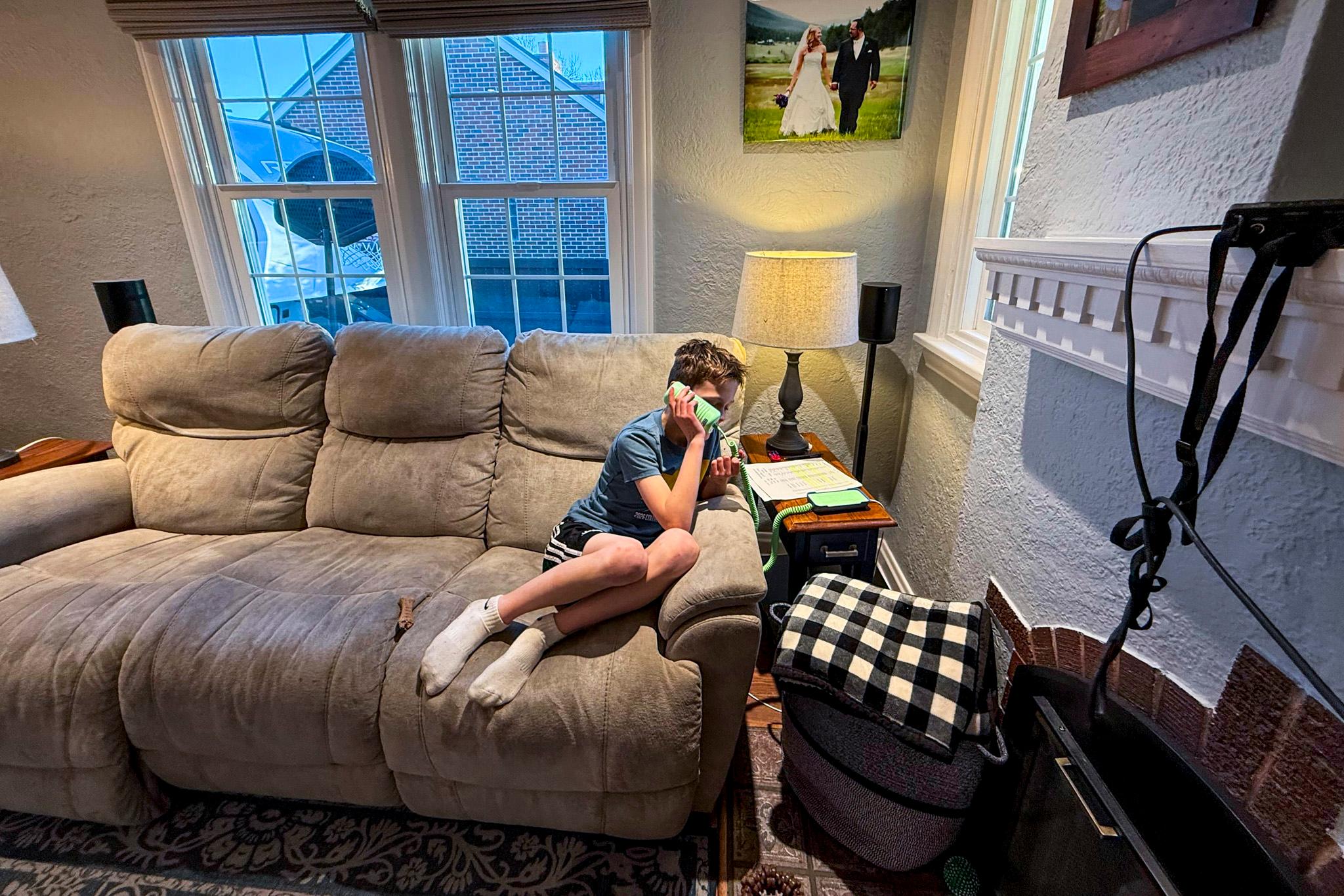 A kid in a t-shirt, shorts and socks sits on a couch, leaning on the arm rest as he talks into a green land-line phone.