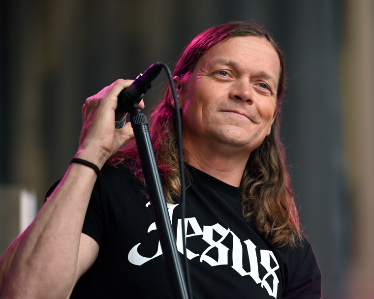 Brad Arnold holds a microphone while smiling. He's wearing a black t-shirt with white letters that say JESUS. 