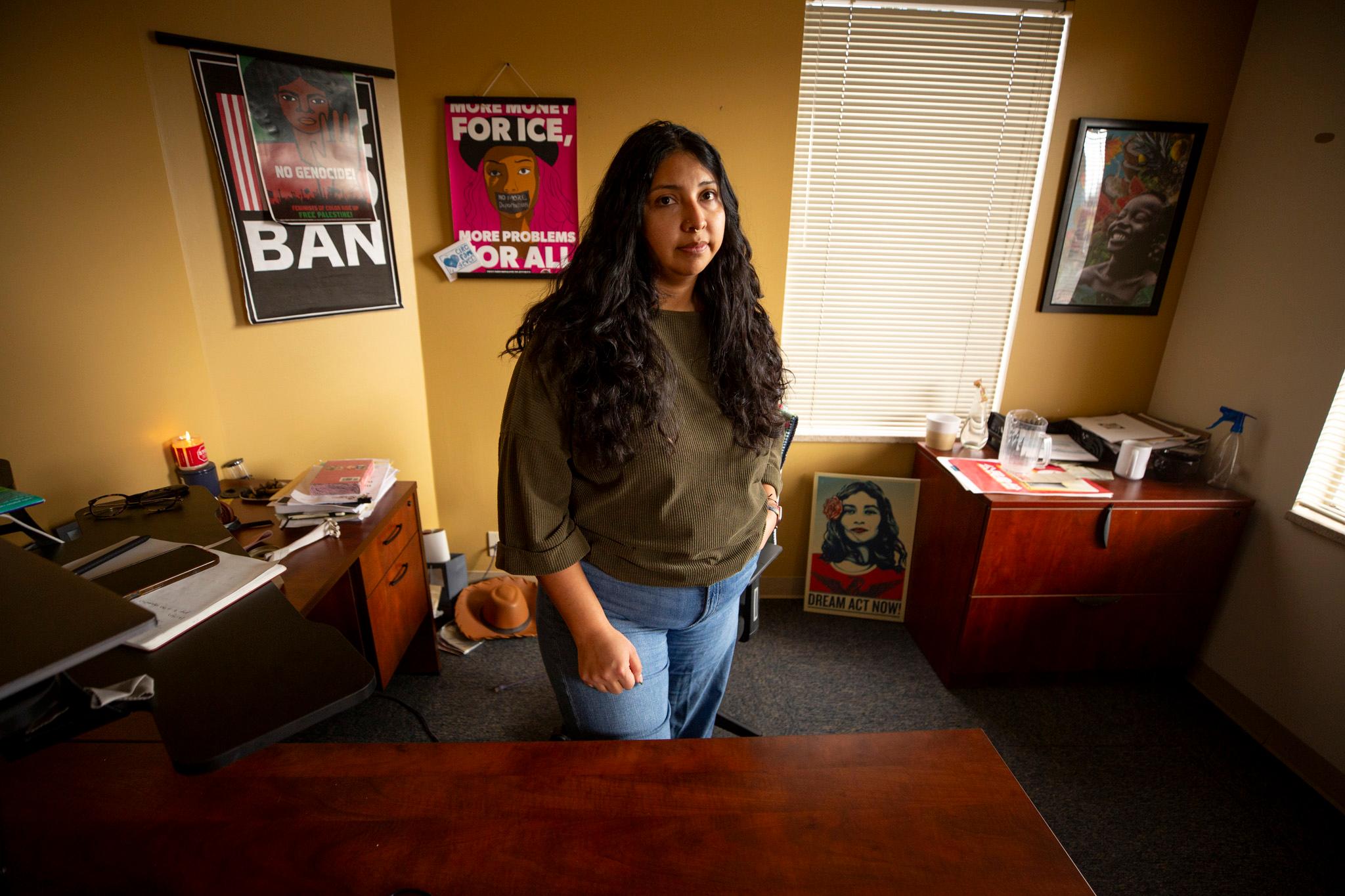 A woman in a brown shirt stands in a warmly lit room, surrounded by posters that read slogans like "More money for ICE, more problems for all."