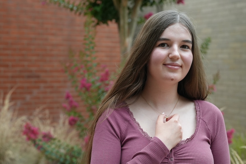 A young woman in front of a garden holds her necklace to her chest. 