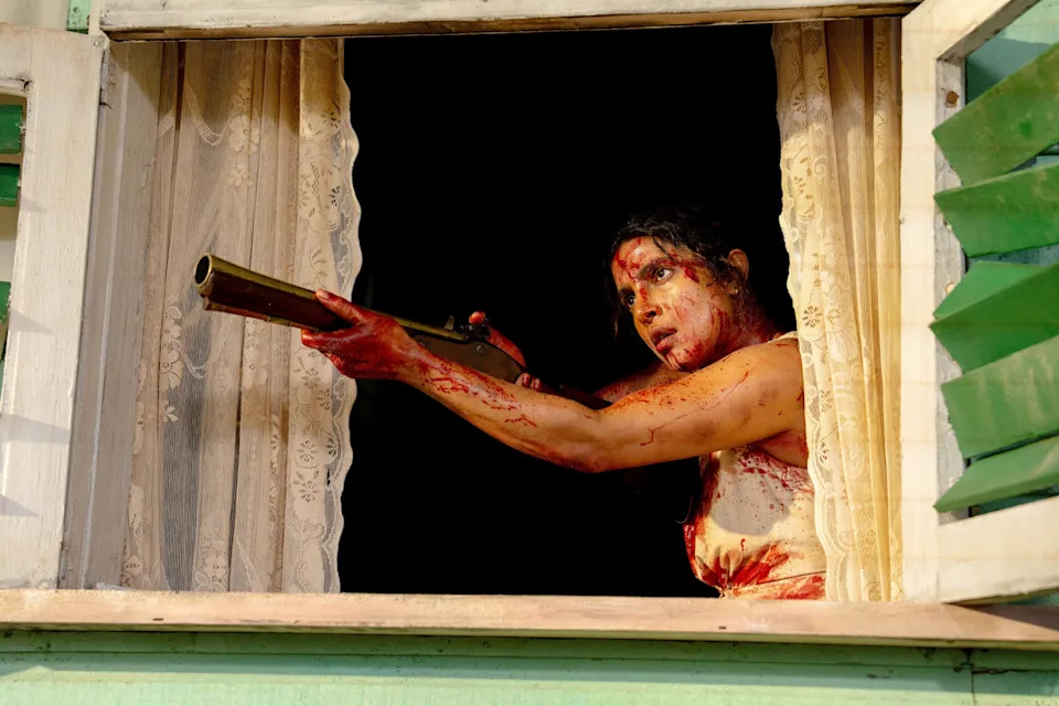 Blood-covered woman aims a shotgun out of a window framed by lace curtains at night.