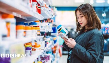 A woman stands in front of store shelves filled with rows of supplement bottles. She is looking at her phone while holding a bottle of supplements.