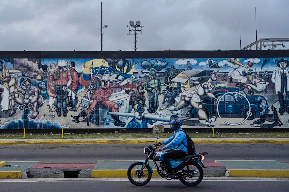 a man on a motorcycle passes a mural on Venezuelan oil topics