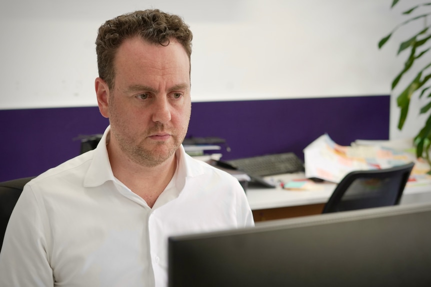 Man with brown hair sits in front of computer in office space.