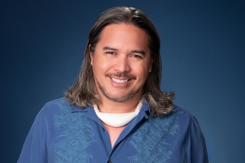 Man in short sleeve button down blue shirt with shoulder length brown hair, moustache, and white necklace. 