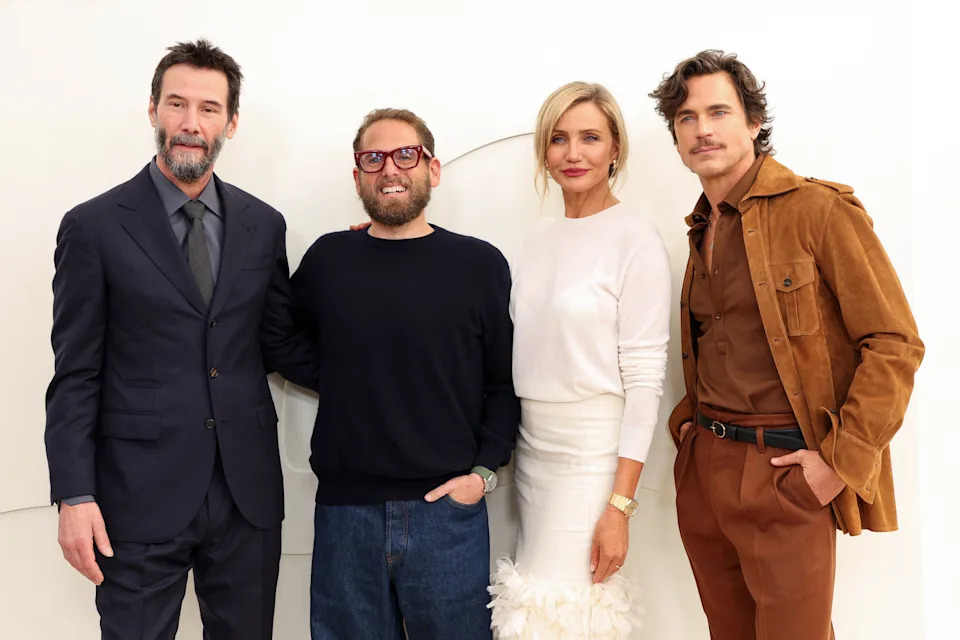 Four people are posing together; one is in a suit, another in a sweater, the third in an elegant outfit, and the fourth in a casual suede jacket