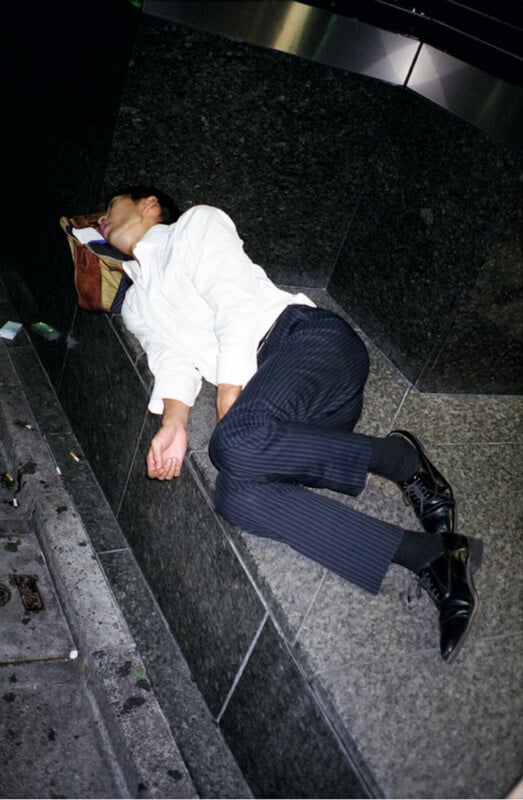 A man wearing a white shirt and dark striped pants sleeps curled up on a stone bench outdoors at night, using a bag as a pillow.