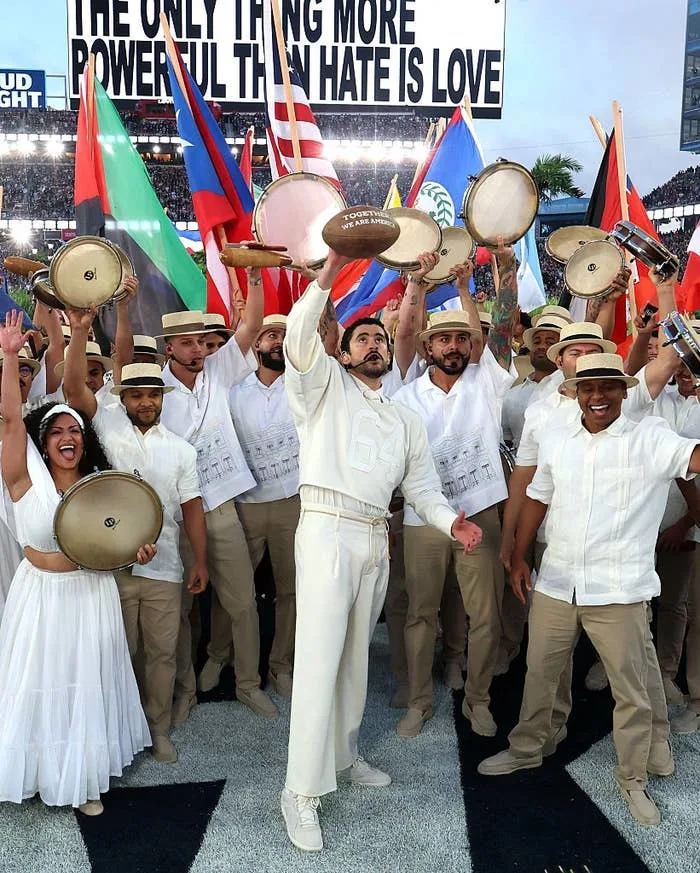 Performers in coordinated attire with tambourines, celebrating on a sports field under a banner reading "The only thing more powerful than hate is love."