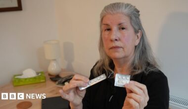 A middle-aged woman with long grey hair holds up two silver-coloured blister packs of round white tablets as she sits in a dining room with white walls. She is wearing a black top and has a serious expression on her face.