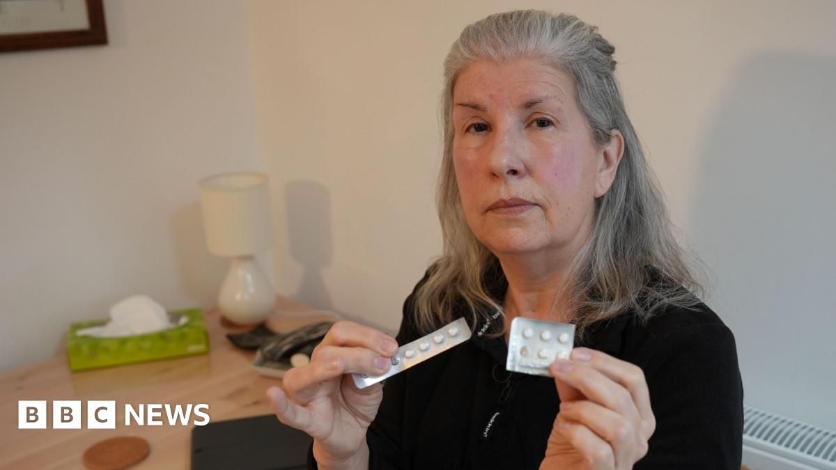 A middle-aged woman with long grey hair holds up two silver-coloured blister packs of round white tablets as she sits in a dining room with white walls. She is wearing a black top and has a serious expression on her face.