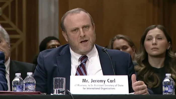 A man in a suit and tie speaks at a formal hearing, seated at a table with a nameplate and water bottles. Audience members listen attentively