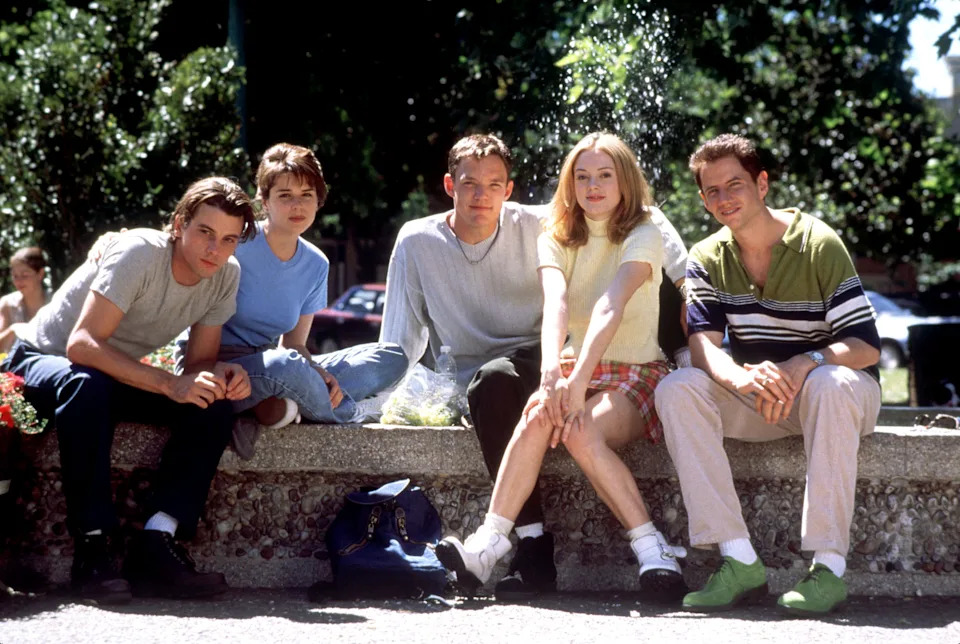 Skeet Ulrich, Neve Campbell, Matthew Lillard, Rose McGowan, and Jamie Kennedy in the original Scream. Five young adults sitting on a stone ledge outdoors with trees behind them, dressed in casual 1990s-style clothing