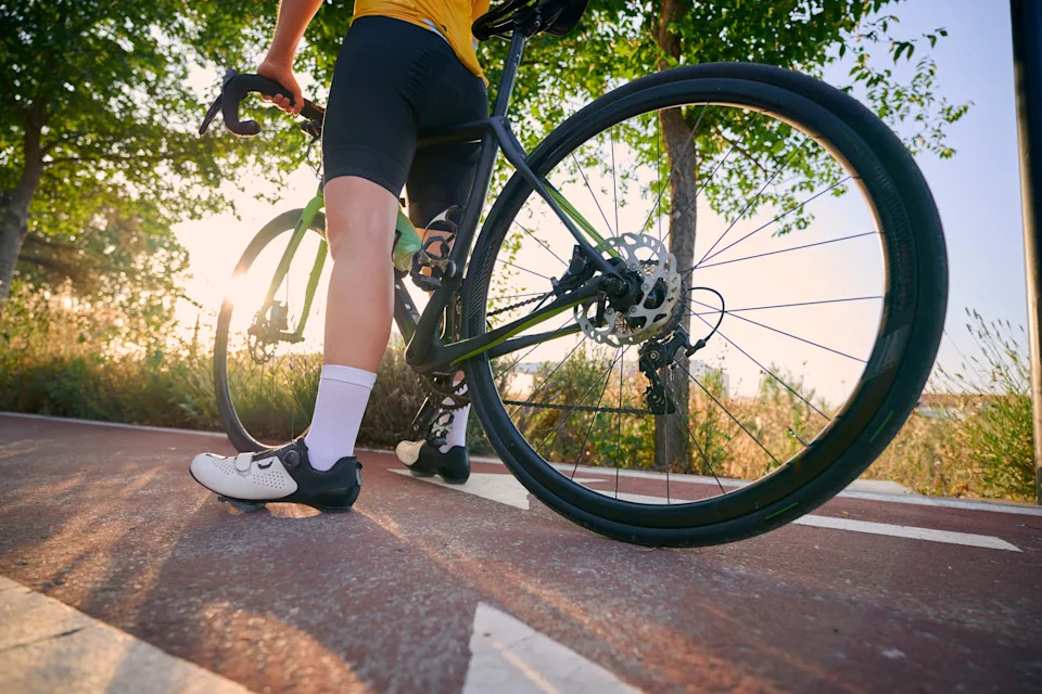 Cyclist standing with bike on a sunlit path, wearing athletic gear and cycling shoes, suggesting an active lifestyle or commute