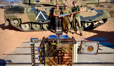 A Flock license plate reader behind a row of shrubs near the entrance of L.A. State Historic Park. A car drives past the park in the background.