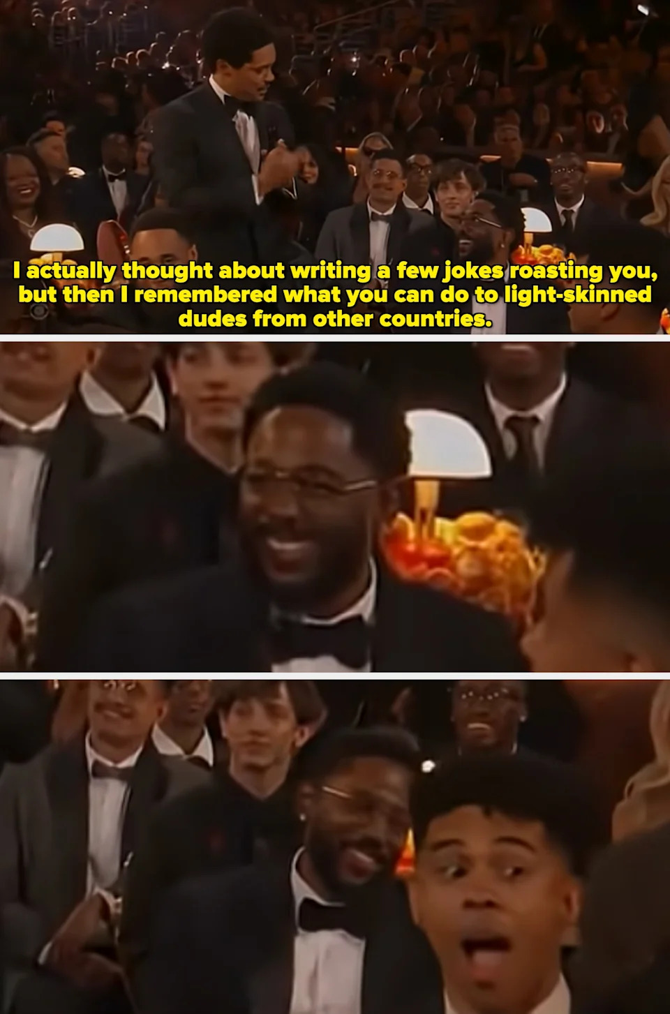 Man in a black tuxedo stands in a crowded audience, holding a microphone, addressing attendees seated around him. Audience members are smiling and engaged