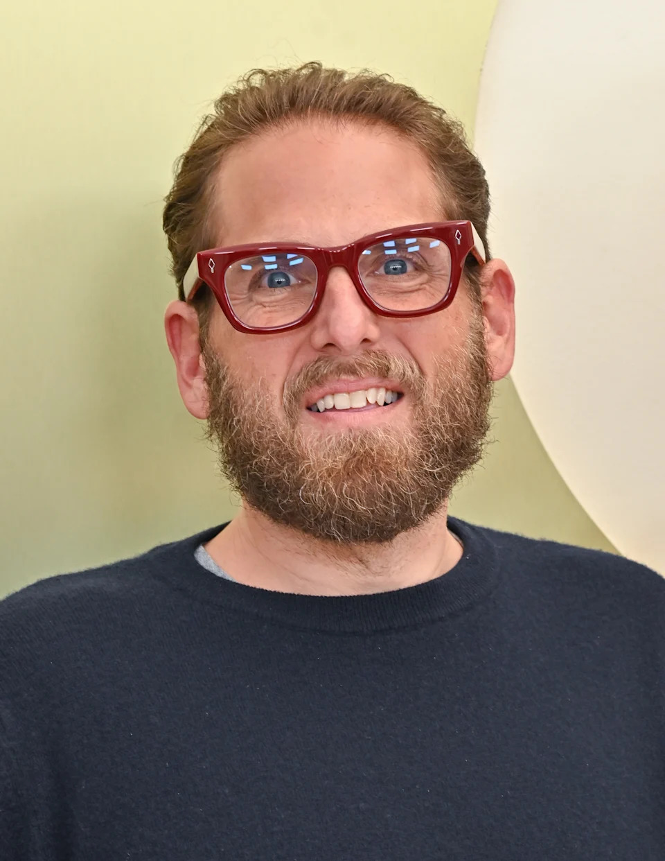 Man with a beard, wearing glasses, smiles while looking at the camera. He is wearing a dark sweater