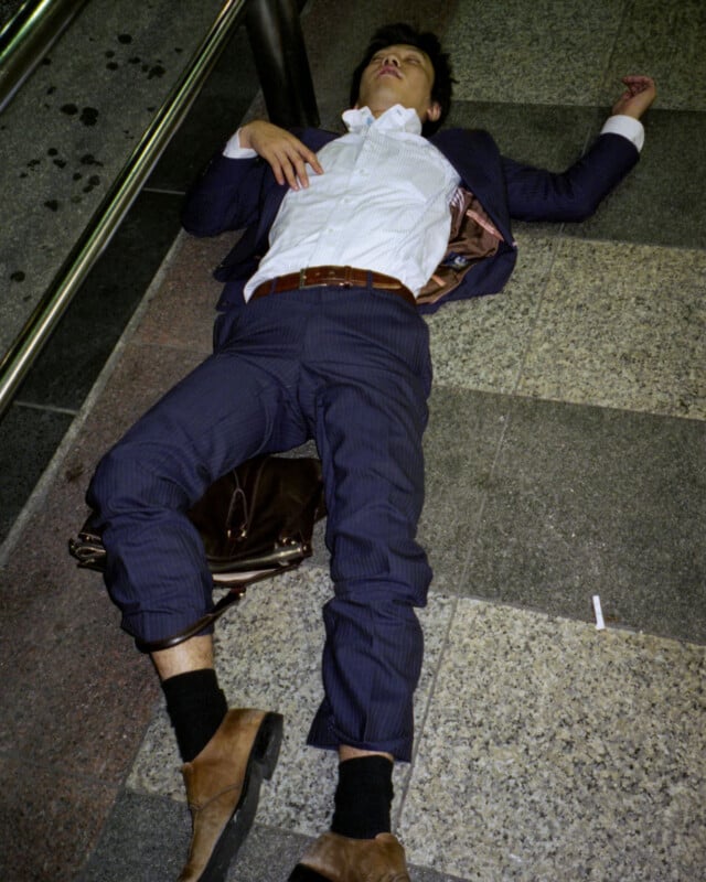 A man in a blue suit lies on his back on a tiled floor with one arm outstretched and eyes closed, appearing unconscious or asleep. A brown bag is beneath him.
