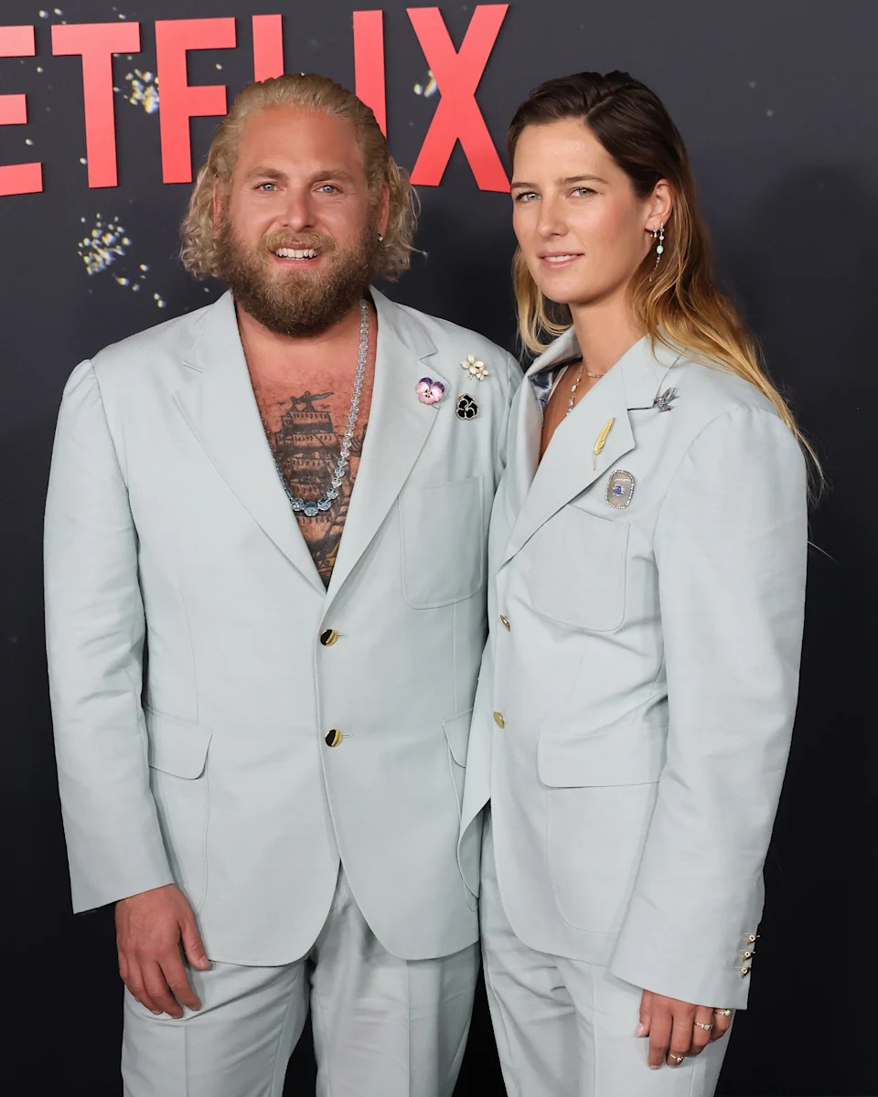 Two people in matching light suits, smiling at a Netflix event backdrop. The person on the left has a beard and visible tattoos