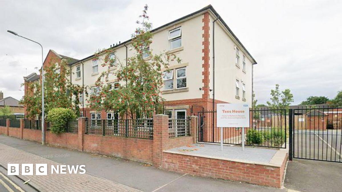 Exterior of a large three-storey building built of brick and partially covered in white rendering. It has black metal gates to the side, leading on to a car park, and a front garden filled with trees and plants