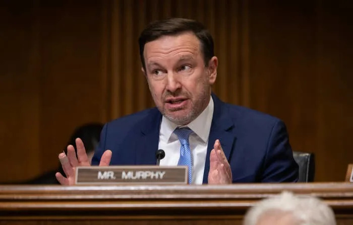 A person in a suit speaks, gesturing with hands, at a formal setting with a nameplate reading "Mr. Murphy" visible. A camera is in the foreground