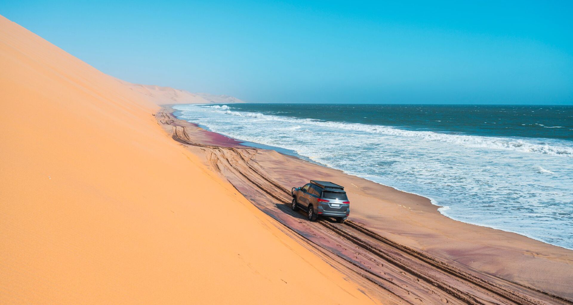 A silver sedan drives on a dark sandy road with a towering desert dune on the left and a blue ocean on the right