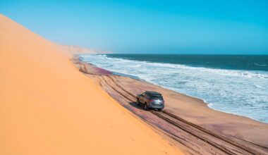 A silver sedan drives on a dark sandy road with a towering desert dune on the left and a blue ocean on the right