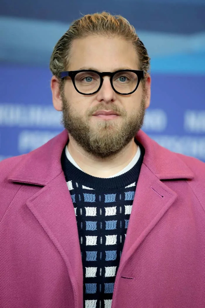 Person with black glasses and a beard wearing a patterned sweater and matching blazer, standing in front of a blurred background
