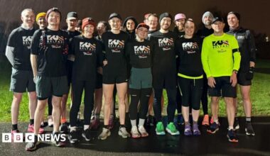 A group of runners standing together. It is raining and night time. Most of them are wearing black T-shirts which say "Run Belfast" on them in white lettering, one man to the front left of the group has the same logo but in black text on a neon yellow running top.