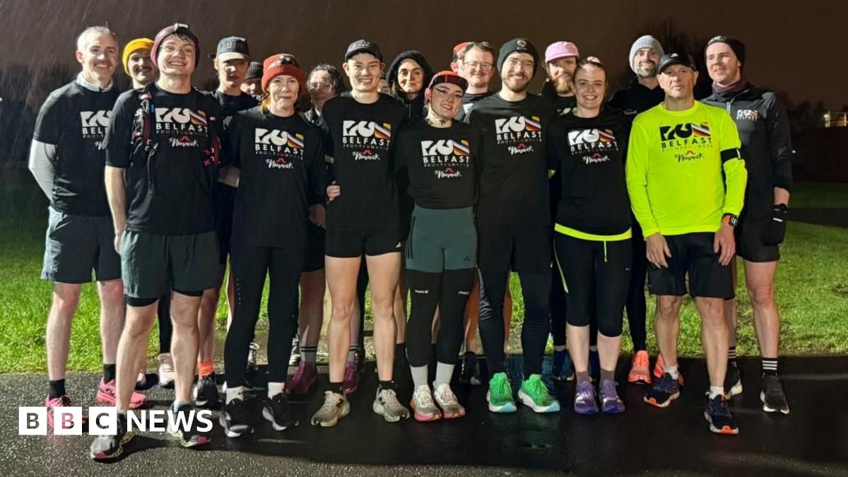 A group of runners standing together. It is raining and night time. Most of them are wearing black T-shirts which say "Run Belfast" on them in white lettering, one man to the front left of the group has the same logo but in black text on a neon yellow running top.