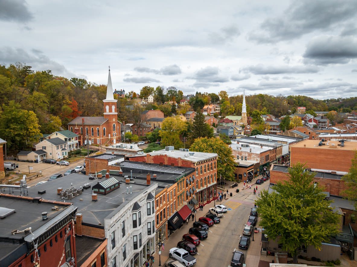 Aerial view of a small town with historic brick buildings, parked cars lining the street, two prominent church steeples, and trees with autumn foliage under a cloudy sky.