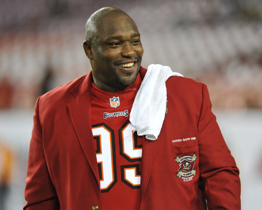 Warren Sapp smiling in a red Tampa Bay Buccaneers blazer with a white towel over his shoulder.