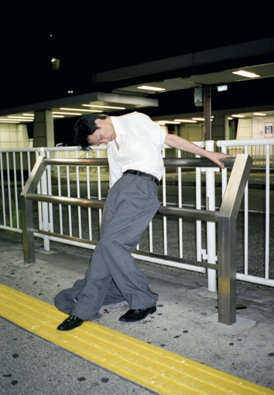 A man in a white shirt and gray trousers leans back on a metal railing at night, appearing tired or unsteady, next to a yellow tactile paving strip on a city sidewalk.