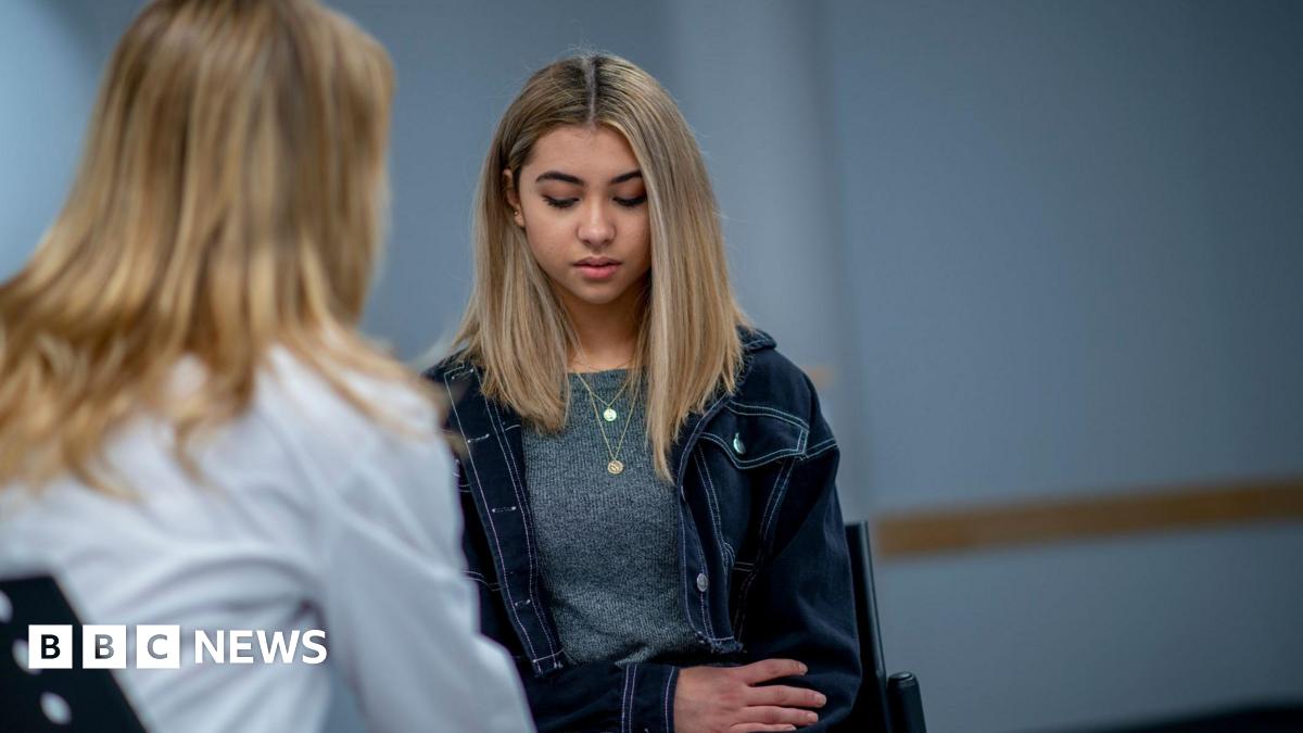 A teenage girl with long blonde hair and wearing a denim jacket and grey T-shirt has her head slightly bowed as she speaks to a woman with long blonde hair and a white shirt who is facing away from the camera.