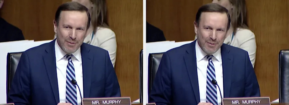 Person speaking at a formal event, wearing a suit and striped tie, seated at a table with a nameplate labeled "Mr. Murphy."