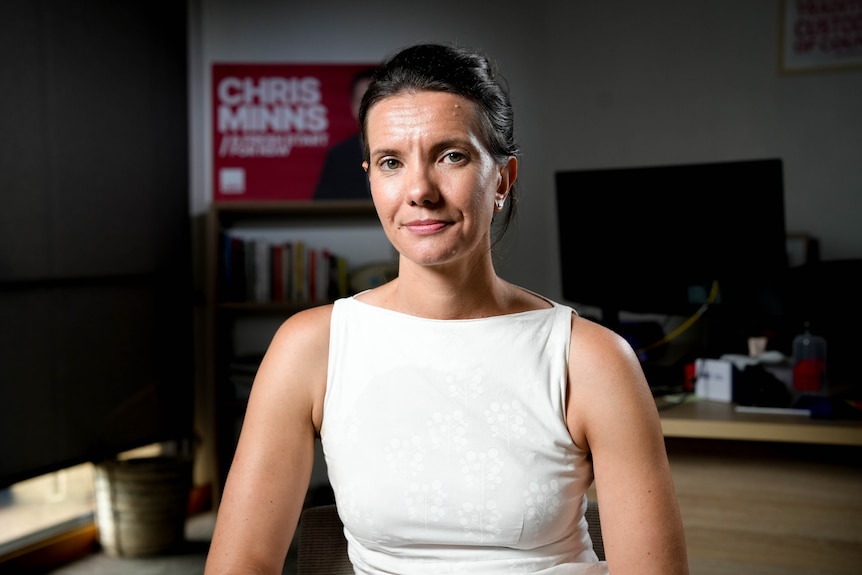 A dark-haired women in sleeveless white dress in an office with a political poster of NSW Premier Chris Minns behind her.