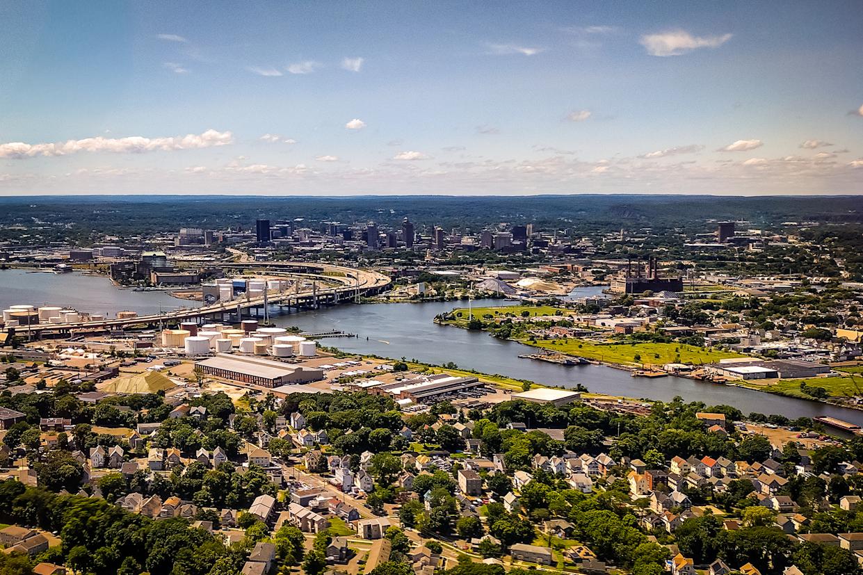 Aerial view of an industrial cityscape with a river, bridges, factories, storage tanks, and residential neighborhoods in the foreground, under a blue sky with scattered clouds.