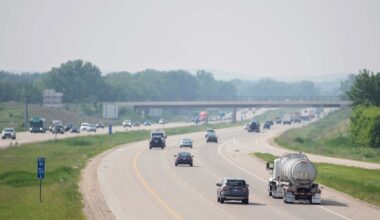 Vehicles, including cars and a tanker truck, travel on a divided highway with light traffic. A bridge crosses the road in the background, and trees line both sides of the highway.
