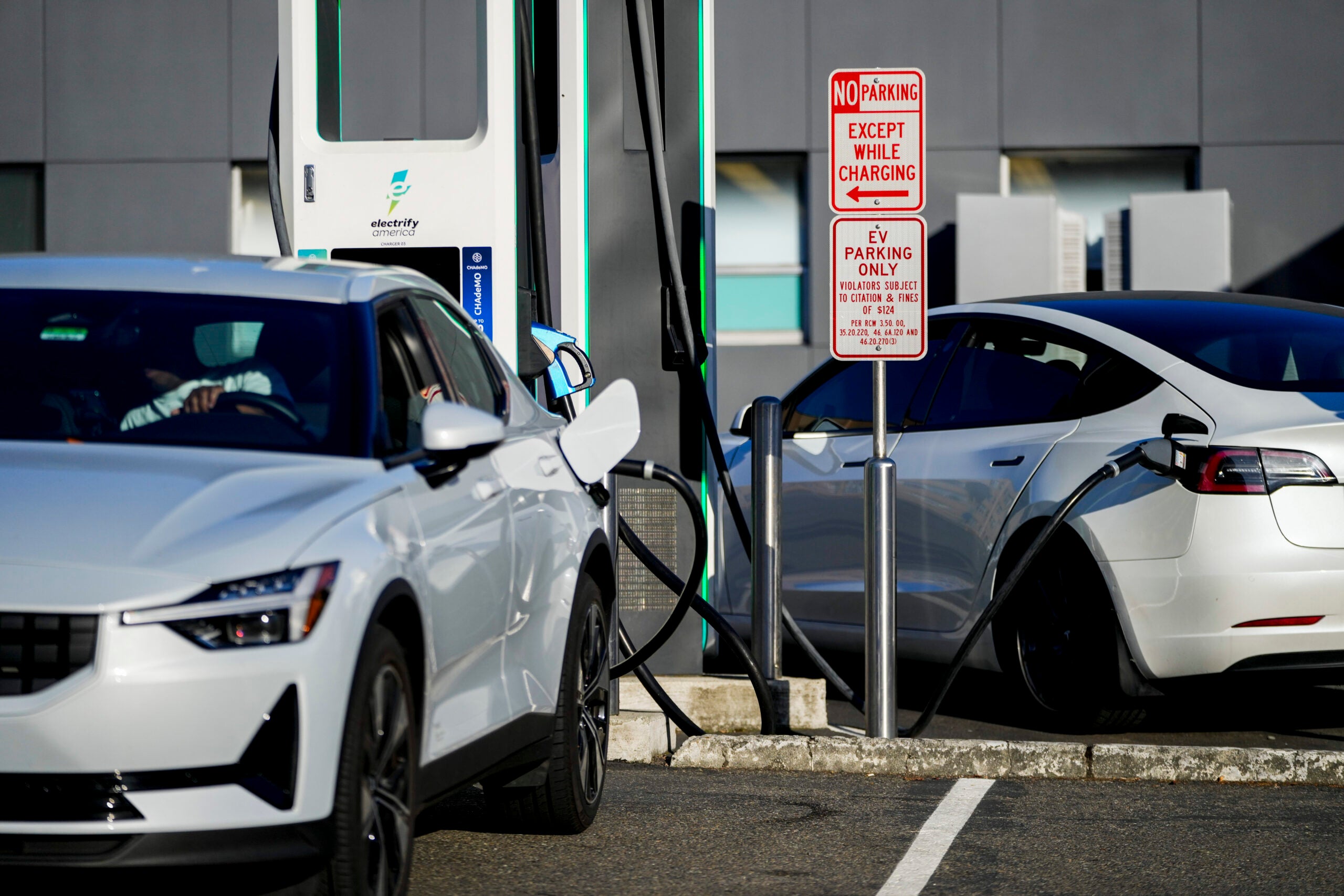Two white electric cars are parked and charging at a charging station in a parking lot with a sign indicating parking is only allowed while charging.