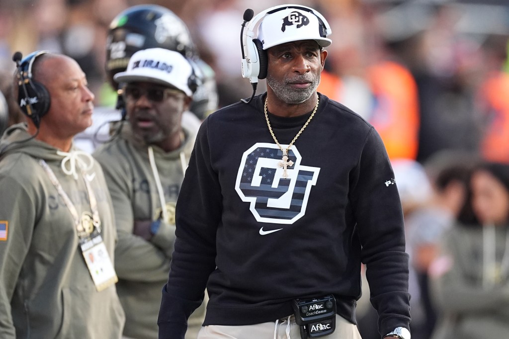 Deion Sanders, head coach for Colorado football, standing on the field wearing a white headset, white cap with the CU logo, and a black CU sweatshirt.