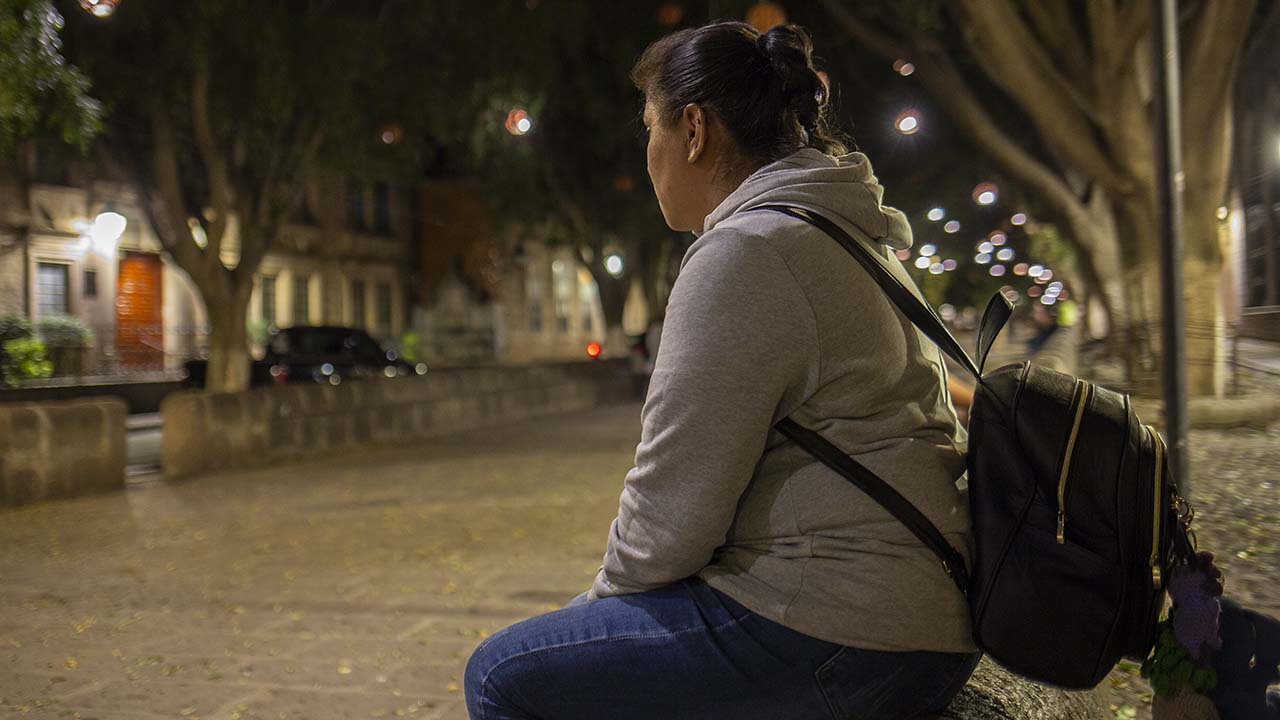 A photo of a woman sitting near a street at night with her back to the camera.