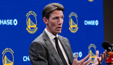 A man in a gray checkered suit and black tie speaks with expressive hand gestures at a table, with Golden State Warriors logos and sponsors behind him.