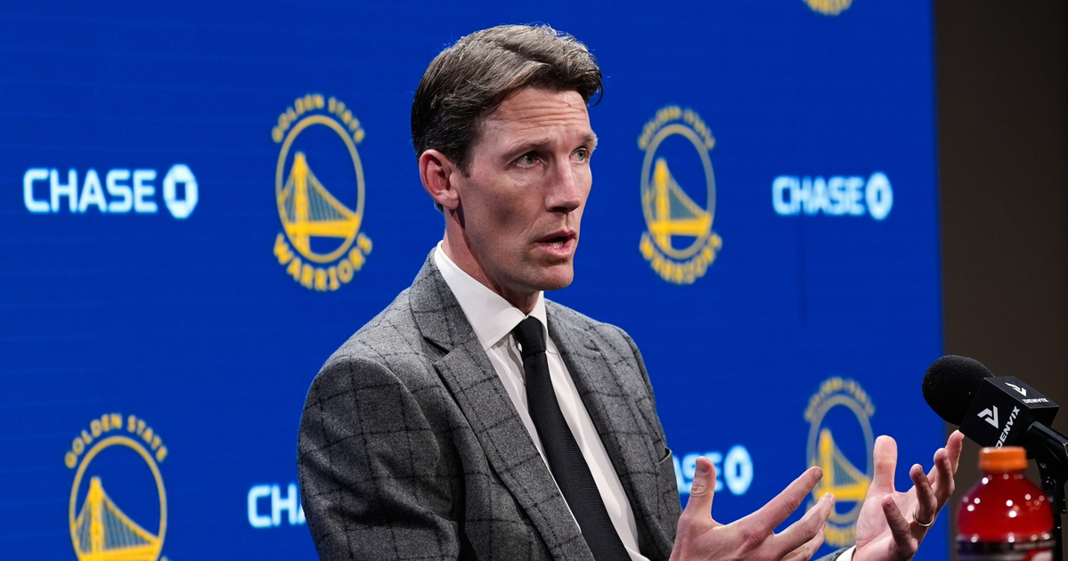A man in a gray checkered suit and black tie speaks with expressive hand gestures at a table, with Golden State Warriors logos and sponsors behind him.