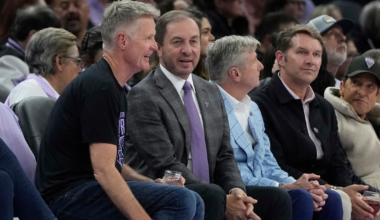 Five men sit closely together in an audience, engaged in conversation, with two holding drinks and wearing casual to business-casual attire.