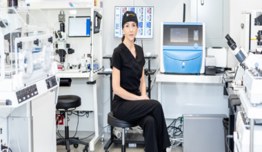 A bobblehead figure of a smiling woman in a white coat and black dress stands on a shelf labeled “Egg Whisperer,” surrounded by books and a white unicorn figurine.