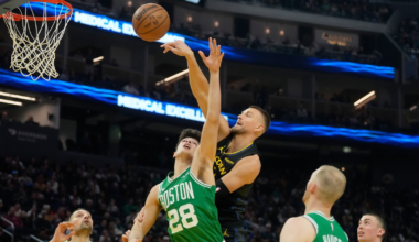 A basketball player in a black jersey jumps to block a shot by a player in a green Boston Celtics jersey near the basket with three other Celtics watching.