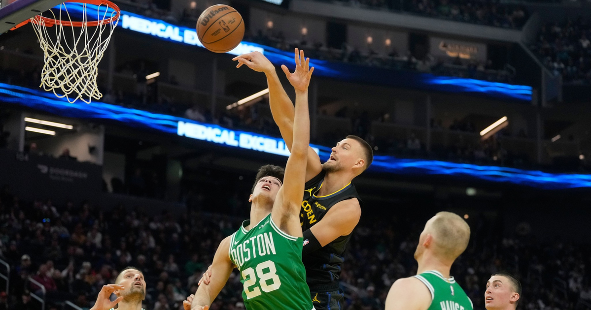 A basketball player in a black jersey jumps to block a shot by a player in a green Boston Celtics jersey near the basket with three other Celtics watching.
