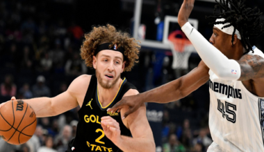 A Golden State player with curly hair drives past a Memphis defender wearing white, both focused intensely as they compete near the basket.